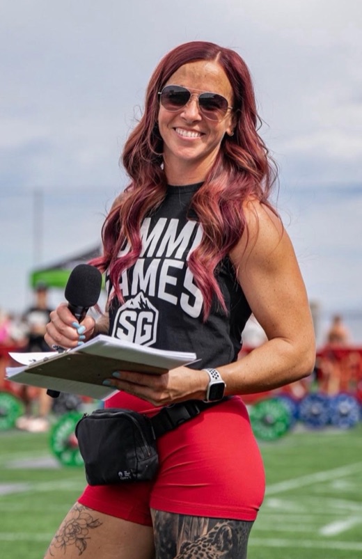 A smiling coach holding a microphone and a clipboard at an outdoor fitness event